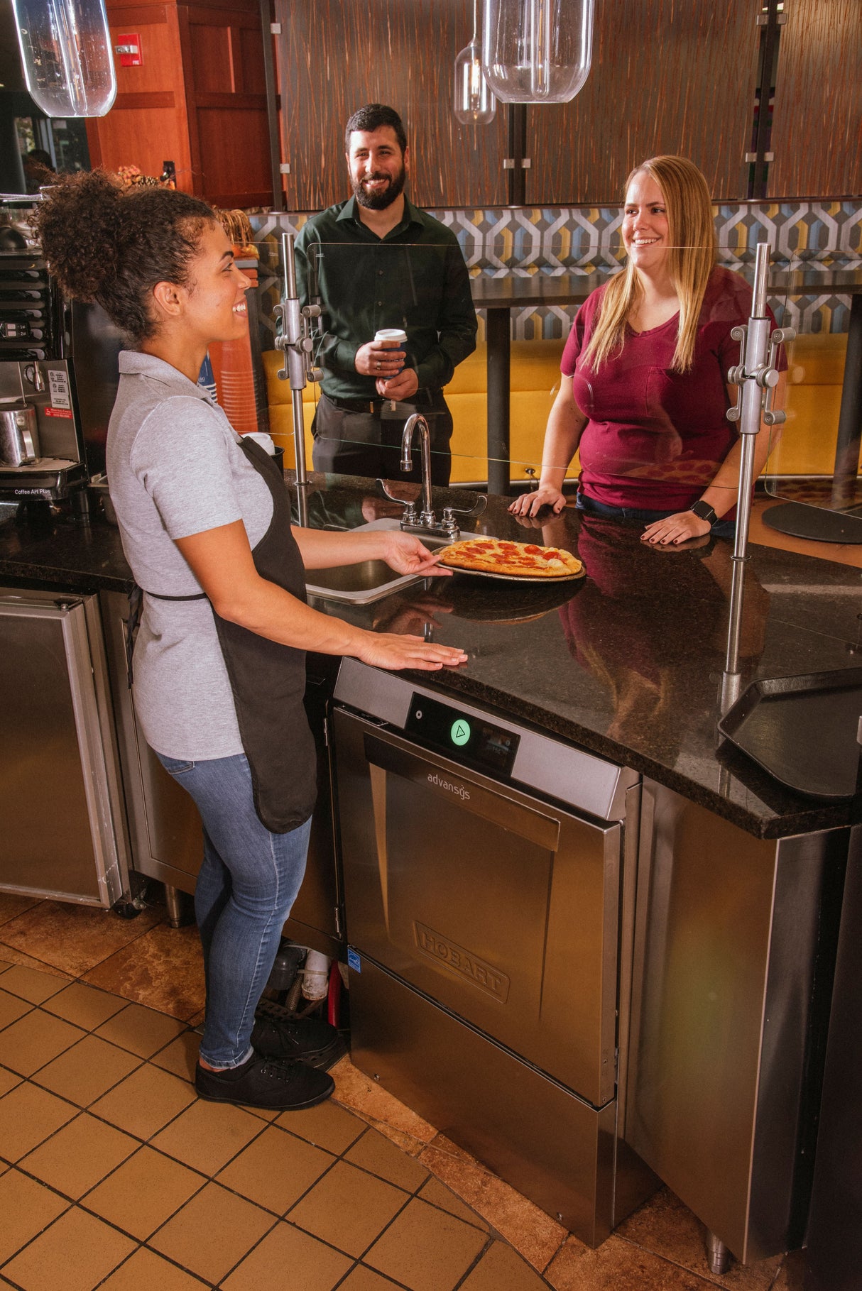 A woman in a gray shirt and black apron stands behind a counter holding a pizza, facing two smiling customers in a modern café with tiled floors, stainless appliances, and an undercounter dishwasher for high temperature sanitizing.