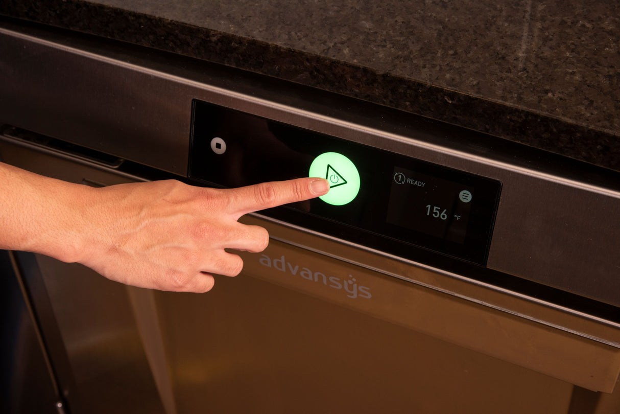 A hand presses a large illuminated green start button on the control panel of an Advansys undercounter commercial dishwasher. The digital display shows READY and 156, all set beneath a dark countertop.
