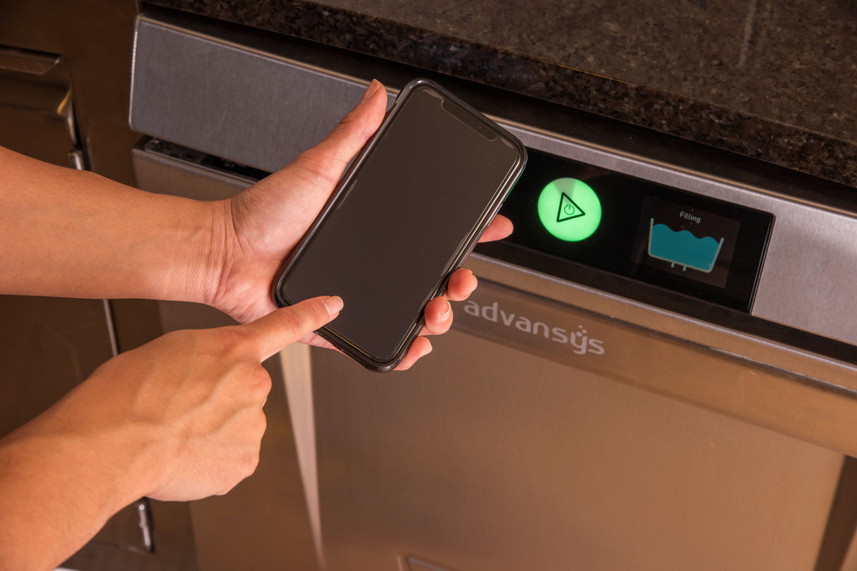 A person holds a smartphone near the control panel of an Advansys undercounter dishwasher. One hand points at the phone while the panel displays a green illuminated button and blue filling icon, highlighting advanced energy recovery features.
