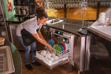 A woman in a gray shirt and black apron loads colorful plates and white bowls into a high temperature sanitizing undercounter dishwasher in a restaurant kitchen. The area is clean, with stainless steel counters, a sink, and a yellow booth in the background.
