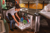 A woman wearing a gray shirt and black apron loads colorful plates, bowls, and cups into an undercounter dishwasher with smart touchscreen controls in a commercial kitchen with stainless steel counters and a clean, organized appearance.