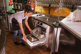 A woman wearing a gray shirt and black apron is kneeling and loading metal trays into a Hobart LXn undercounter dishwasher in a commercial kitchen. Stainless steel counters surround her as she smiles, focused on high temperature sanitizing.
