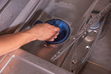 A hand is cleaning the round blue filter inside the bottom of an undercounter dishwasher. The metal interior, spinning spray arm, and perforated base highlight routine maintenance for optimal high temperature sanitizing performance.