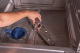 A hand uses pliers to loosen the central bolt of a metal spray arm inside an open undercounter dishwasher, near the filter basket and drain area, showing the process of removing dishwasher parts for cleaning or maintenance.