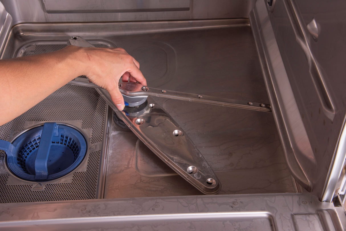 A person’s hand removes or installs the lower spray arm inside an empty stainless steel Hobart LXn Dishwasher. The blue filter basket and fine mesh filter are visible on the left side of the undercounter dishwasher’s clean, metallic base.