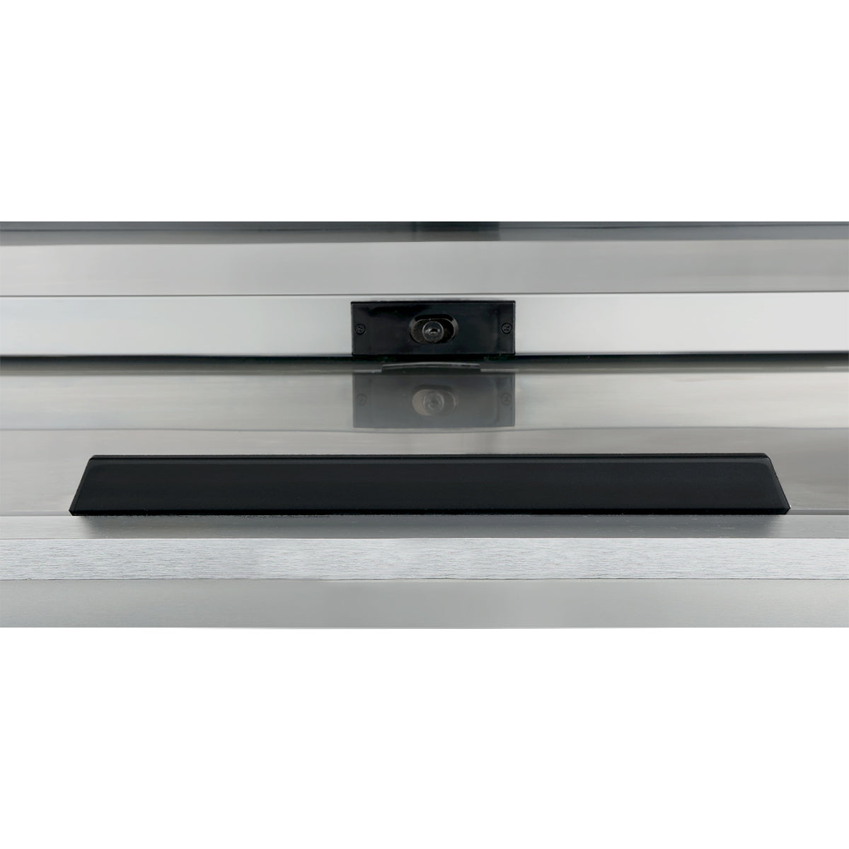 A rectangular black object sits centered on a stainless steel surface in front of a reflective glass panel. Behind the glass, a small dark rectangle with a round feature, possibly an Empura refrigeration sensor, is visible. The scene is minimal and industrial.