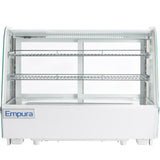 A white Empura refrigerated display case sits on the counter, featuring two glass shelves, clear curved glass front, and interior LED lighting. This countertop display case is empty, with a blue Empura logo on the bottom left corner.