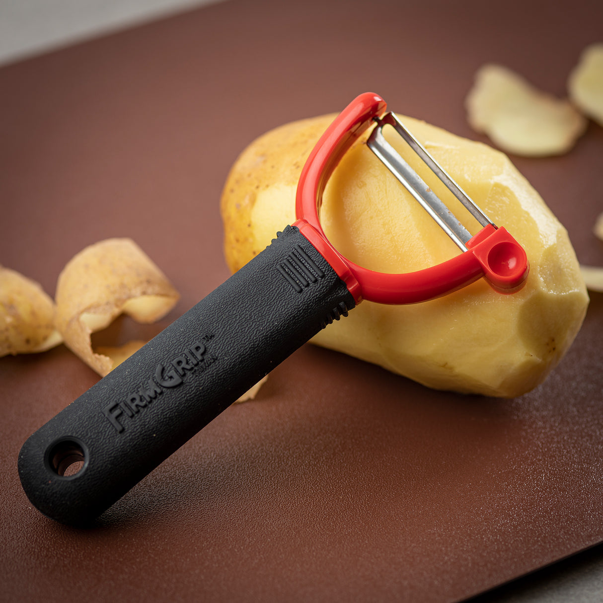 A black and red Y peeler with an ergonomic handle rests on a partially peeled potato, with potato skins scattered on a brown surface in the background.