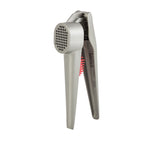 A stainless steel garlic press with an ergonomic handle and perforated chamber stands upright. A red cleaning tool with bristles is partially visible, stored inside one of the handles. The background is plain white.