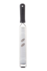 A Microplane Grater with an ergonomic handle is shown vertically on a white background. Two whole nutmeg seeds rest on its sharp, small holes for fine grating. The rectangular, flat grater is also dishwasher safe for easy cleaning.