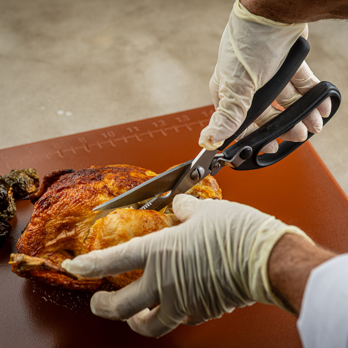 A person wearing white gloves uses stainless steel poultry shears with a firm grip to cut into a roasted chicken on a brown cutting board. The scene is set in a kitchen, with part of the chicken already separated.