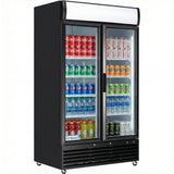 A black refrigerated merchandiser with glass swing doors displays Coca-Cola, Sprite, Pepsi, and Fanta cans on shelves, while bottled juice, water, and Monster energy drinks sit below. This commercial refrigerator features two doors and interior lighting.