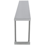 A long, narrow stainless steel table is shown from the short end, emphasizing its length. The smooth top and four cylindrical metal legs feature a sturdy table-mounted horizontal support bar. The background is plain and white.