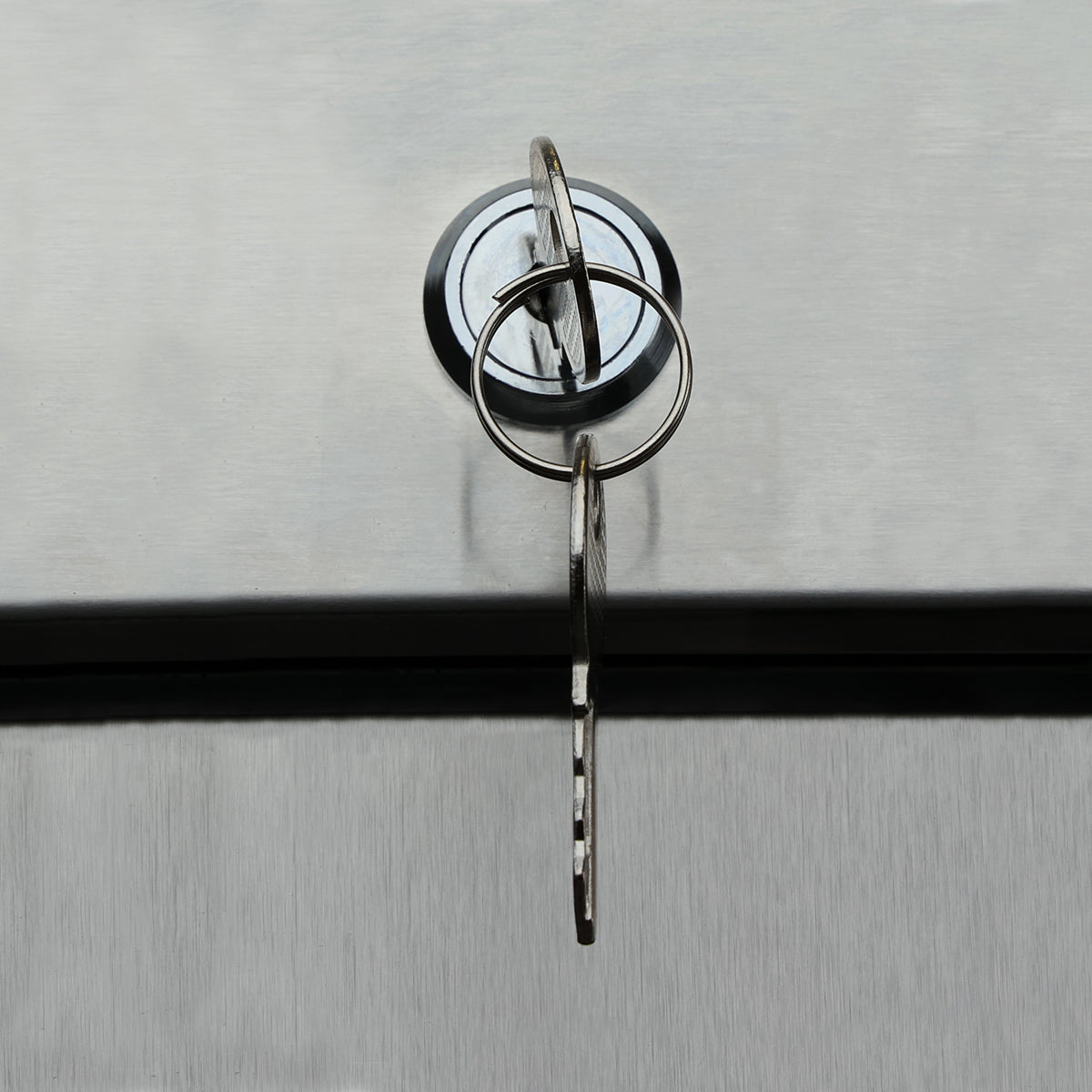 A close-up of a metallic key inserted into a circular lock on the brushed stainless steel surface of an Empura Refrigeration reach-in freezer. The key, attached to a metal keyring, casts faint shadows on the reflective background.