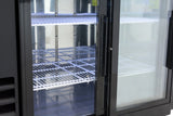 A commercial refrigerator with sliding glass doors stands open, revealing empty white wire shelves inside. The shelves are clean and evenly spaced, and the interior is brightly lit. No products or items are visible inside this commercial refrigeration unit.