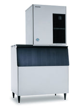 A large, commercial Hoshizaki ice maker with a rectangular silver body, black vent on the top right, and a black front bin lid. The unit stands on four black legs and has a blue circular logo on the front lower section.