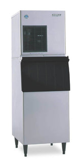 A Hoshizaki stainless steel ice maker with a black front panel stands on four legs. The upper section has a vent and control panel, while the lower features a pull-down door for dispensing cubelet-style ice. Its design is simple and industrial.