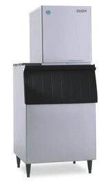 A Hoshizaki commercial Ice Maker featuring a tall, silver rectangular top unit and large black-fronted storage bin, stands on four legs. The flake-style machine boasts clean lines and a durable stainless steel finish for a boxy, modern look.