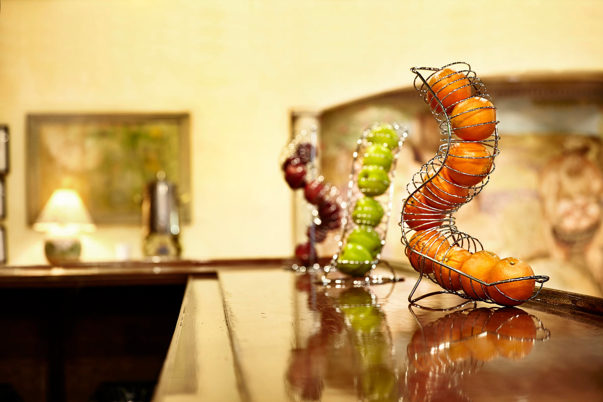 A chrome plated TableCraft fruit basket holds oranges, green apples, and red apples on a shiny wooden counter. The softly blurred background features framed art, a lamp, and warm lighting for a cozy, inviting atmosphere.
