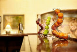 A chrome plated TableCraft fruit basket holds oranges, green apples, and red apples on a shiny wooden counter. The softly blurred background features framed art, a lamp, and warm lighting for a cozy, inviting atmosphere.
