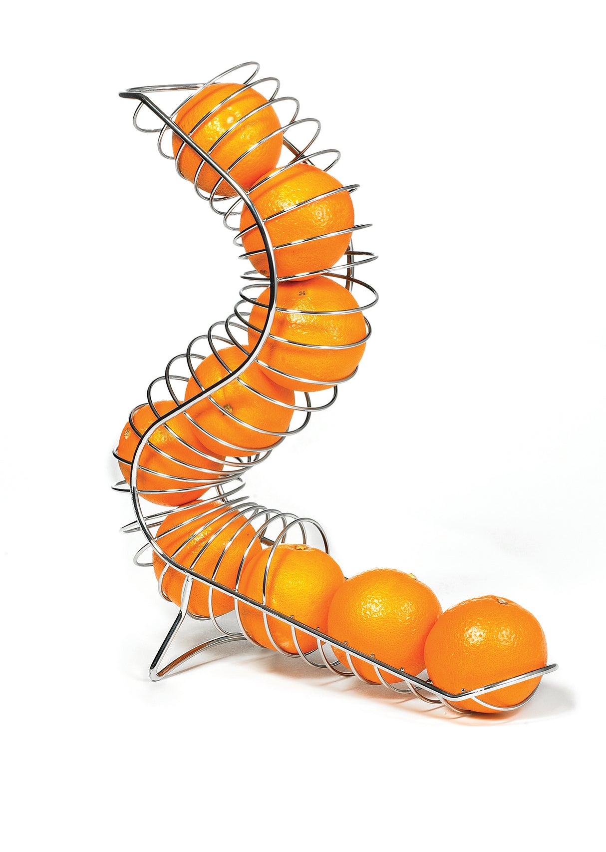 A modern, spiral-shaped Chrome Plated Fruit Basket by TableCraft is filled with bright oranges. The S-shaped holder balances on three legs against a plain white background, with the oranges neatly lined up inside the curved wire structure.