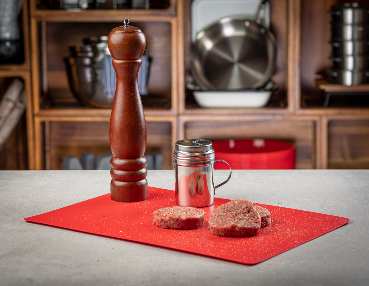 A wooden pepper grinder, a metal shaker, and three raw hamburger patties sit on a red polyethylene cutting mat, lightly sprinkled with seasoning. In the background, kitchen shelves hold pots, pans, and dishes.