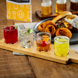 A wood paddle flight holds four colorful cocktails, each garnished with fruit or herbs. Behind them are a plate of onion rings and a burger, and three small glasses of beer. The setting is a wooden table with a yellow menu in the background.