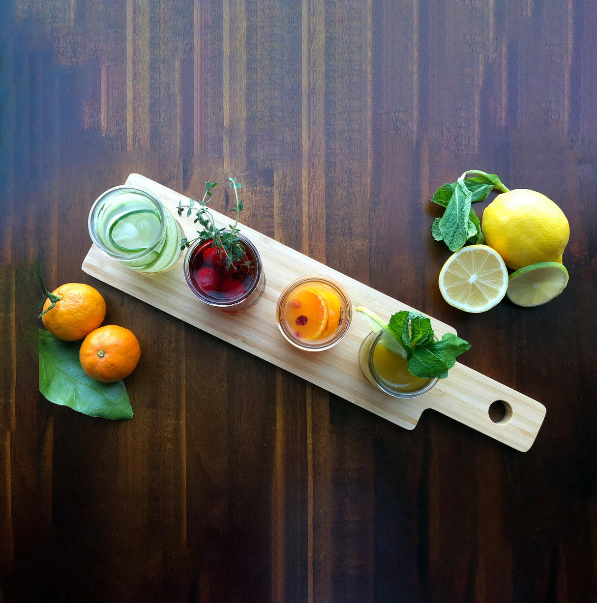 A TableCraft FLIGHT1 bamboo flight board holds four glass jars filled with colorful drinks, each garnished with herbs and fruit. Around the wood paddle flight are tangerines, fresh mint, and sliced lemons on a dark wooden table.