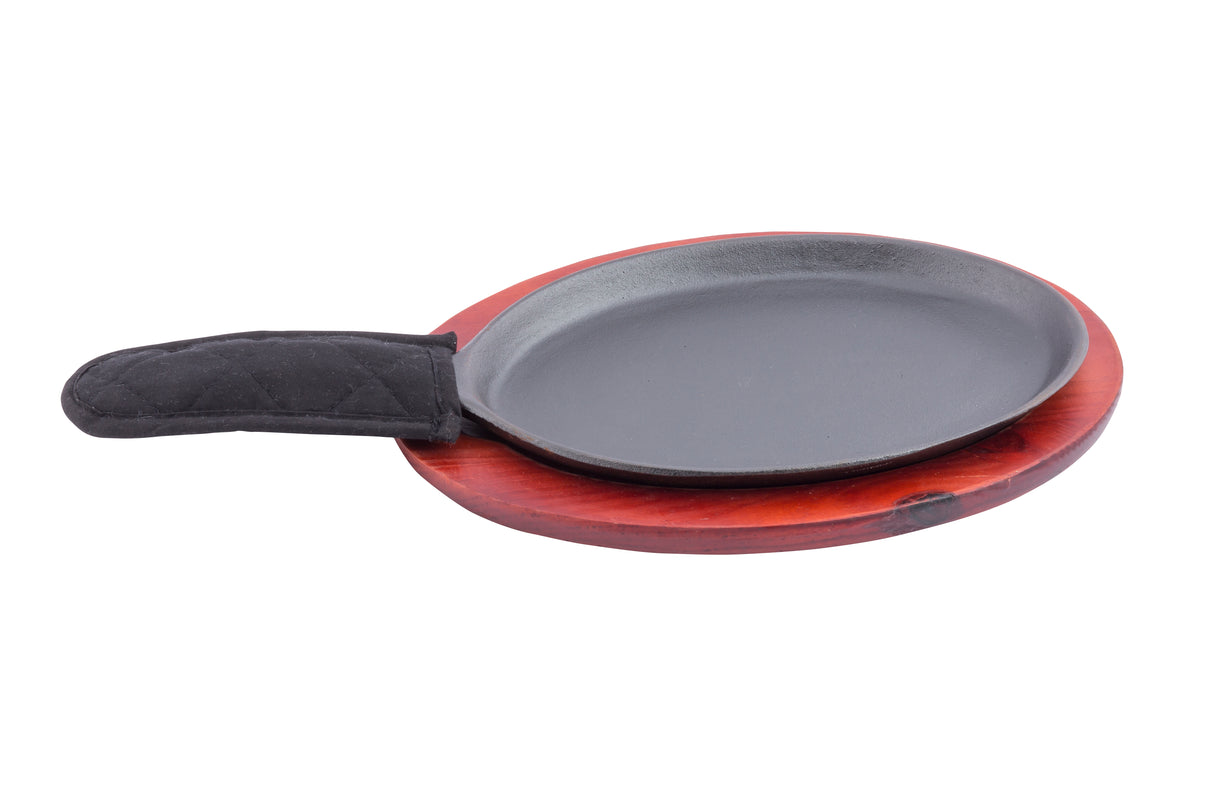 A black, oval-shaped Cast Iron Sizzle Platter with a quilted black fabric handle cover sits on a round, reddish-brown wooden base, isolated on a white background.