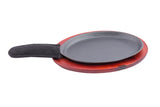 A black, oval-shaped Cast Iron Sizzle Platter with a quilted black fabric handle cover sits on a round, reddish-brown wooden base, isolated on a white background.