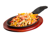 A sizzling Cast Iron Sizzle Platter filled with cooked chicken strips, colorful bell peppers, onions, and herbs sits on a wooden serving board. The handle is covered with a black protective sleeve, set against a white background.