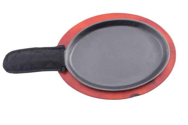 A round black cast iron sizzle platter with a black quilted fabric handle cover rests on a circular reddish-brown wooden trivet, shown from above on a white background. Perfect for serving sizzling fajitas, this is quality TableCraft Products craftsmanship.