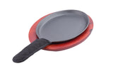 A black cast iron sizzle platter with a fabric handle cover sits on a reddish-brown wooden serving base. The TableCraft Products skillet is empty and both items are placed on a white background.