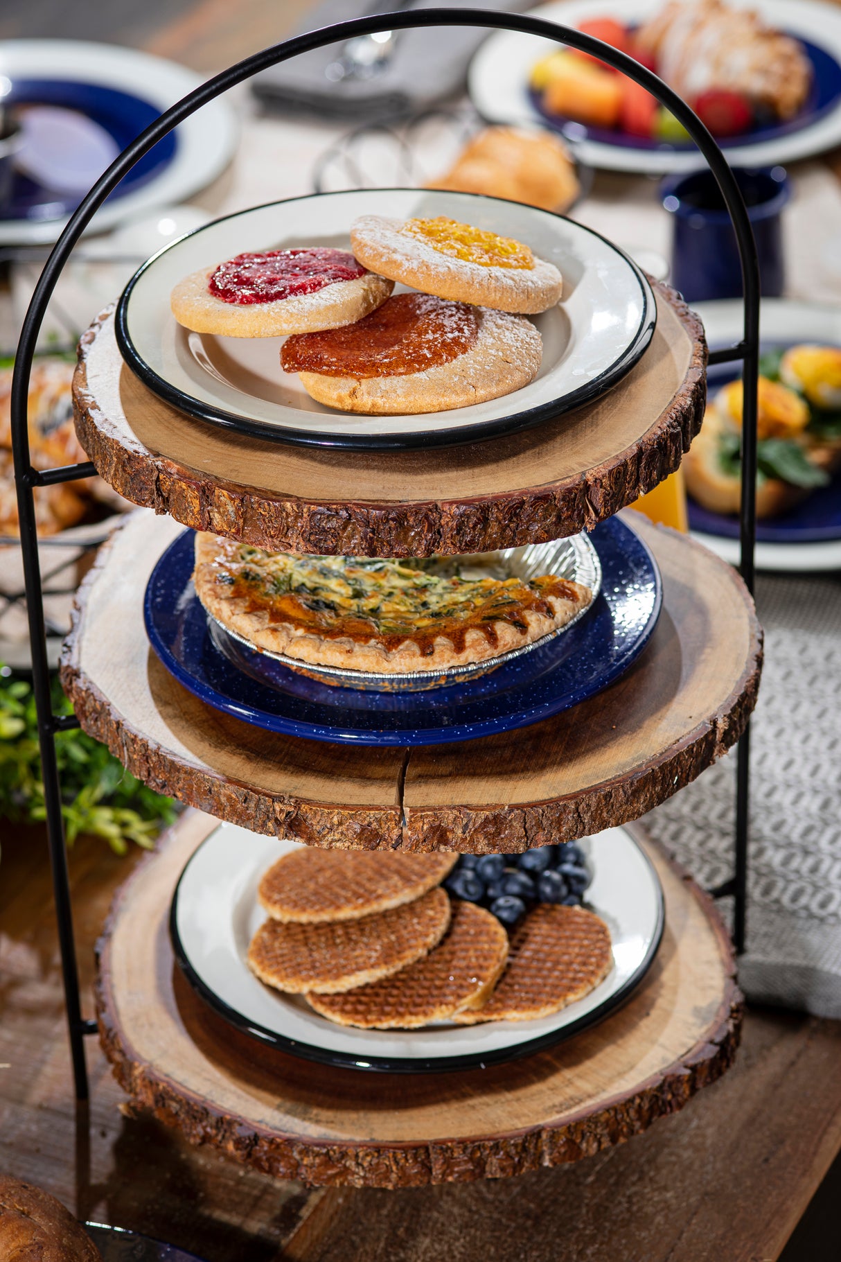 A 3-tier wooden stand, reminiscent of a Fajita Rack, holds assorted pastries: cookies topped with jam, a vegetable quiche, and stroopwafels with blueberries. Plates blur in the background, enhancing the cozy brunch scene.