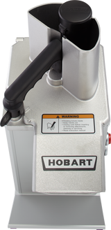 Top view of a Hobart stainless steel food processor with a large handle, black safety lid, and warning label about sharp blades. The metallic machine has an industrial design and features continuous feed for efficient processing.