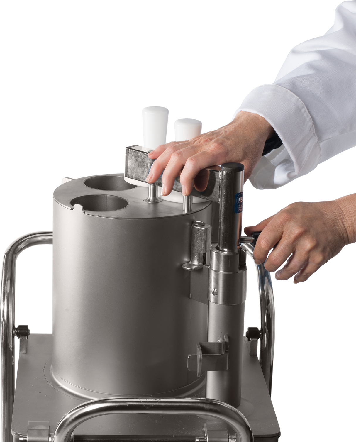 A person in a white lab coat operates a continuous-feed food processor with two white handles and a metal lever. The cylindrical stainless steel machine sits on a metal cart against a plain white background; only the hands and forearms are visible.