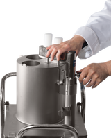A person in a white lab coat operates a continuous-feed food processor with two white handles and a metal lever. The cylindrical stainless steel machine sits on a metal cart against a plain white background; only the hands and forearms are visible.