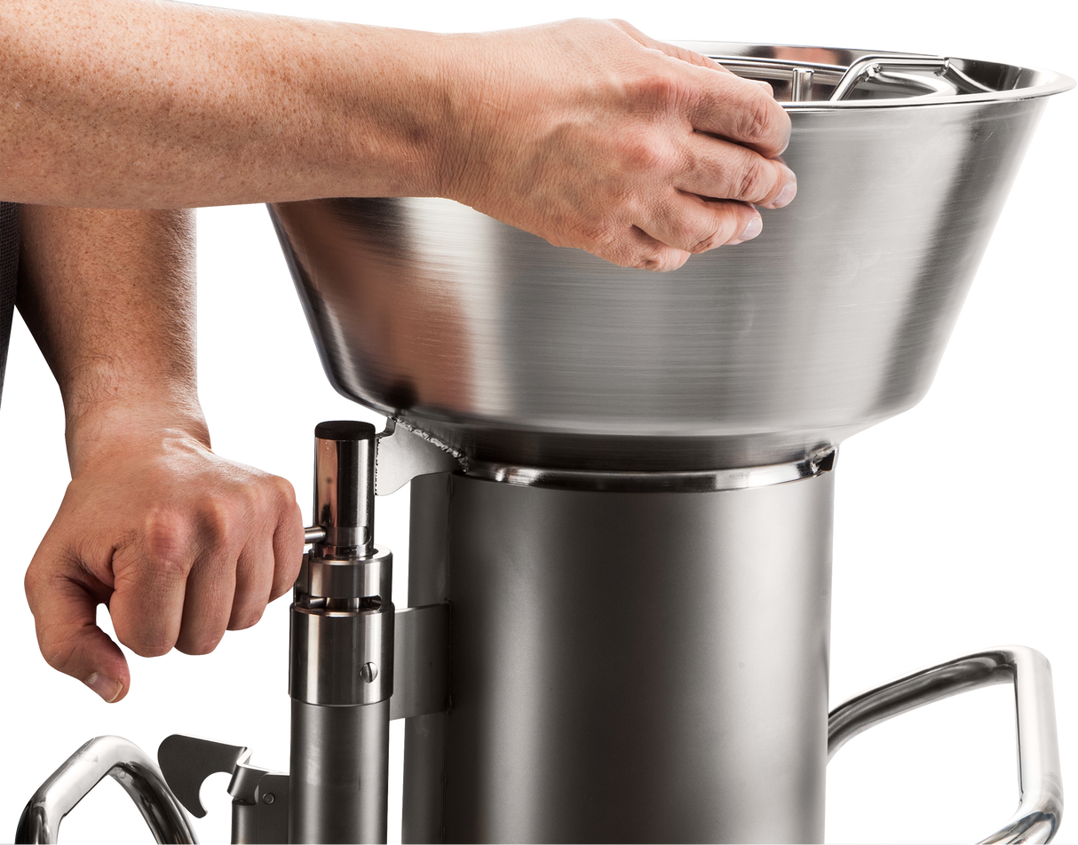A close-up of two hands operating a high production food processor sausage stuffer. One hand turns the handle as the other steadies the large metal hopper atop the cylindrical main body, all against a plain white background.