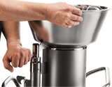 A close-up of two hands operating a high production food processor sausage stuffer. One hand turns the handle as the other steadies the large metal hopper atop the cylindrical main body, all against a plain white background.