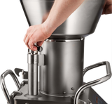 A person’s hand adjusts a black knob on a Hobart FP400I Continuous-Feed Food Processor, a large stainless steel machine with a funnel-shaped top and sturdy handles, capable of processing up to 132 lb/minute with its robust, industrial design.