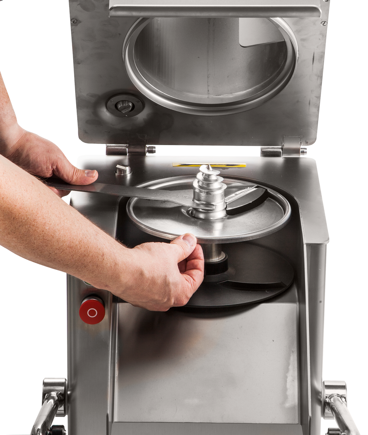 A person’s hands install a circular metal blade into a high production food processor with a stainless steel body. The machines open lid reveals its interior and round blade compartment, while a red button is visible on the side.
