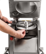 A person’s hands install a circular metal blade into a high production food processor with a stainless steel body. The machines open lid reveals its interior and round blade compartment, while a red button is visible on the side.