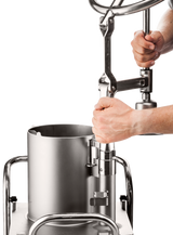 A person’s hands operate a large, stainless steel sausage stuffer, turning a crank handle above a cylindrical container. Similar to the efficiency of the Hobart FP400I Continuous-Feed Food Processor, this machine is set on white with shiny metal rails.
