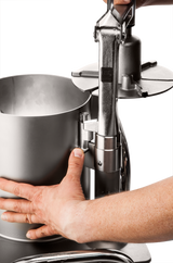 Close-up of two hands operating a high production food processor, with one hand steadying the cylindrical container while the other pulls down a lever attached to a disc, likely for slicing or pressing food inside.