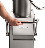 A hand grips a metal handle on a stainless steel Hobart FP400I Continuous-Feed Food Processor. The 132 lb/minute machine displays the Hobart logo and a yellow warning label. Part of a person’s arm and gray shirt are visible on the left.