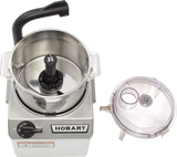 A top view of a silver Hobart food processor with an open stainless steel 4 qt bowl, revealing a sharp S-blade. The clear plastic lid with feed tube and cap rests to the side, while the control knob is set to off/pulse.