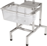 A transparent plastic infant bassinet rests on a sturdy, rectangular stainless steel frame with an FP100 mount at one end. The frame features rubber pads on the feet for stability. The bassinet is empty, set against a plain white background.