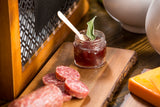 A small glass tasting jar of red jam with a wooden spoon and green herb sits on a wooden board beside salami slices and orange cheese; the rustic setting suggests an artisanal charcuterie arrangement.