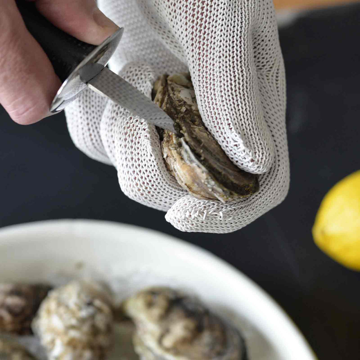 A hand wearing a cut resistant glove holds an oyster while using an oyster knife to pry it open. Nearby, a bowl of oysters and a yellow lemon set the scene for oyster shucking preparation, prioritizing safety with the protective glove.