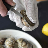 A hand wearing a cut resistant glove holds an oyster while using an oyster knife to pry it open. Nearby, a bowl of oysters and a yellow lemon set the scene for oyster shucking preparation, prioritizing safety with the protective glove.